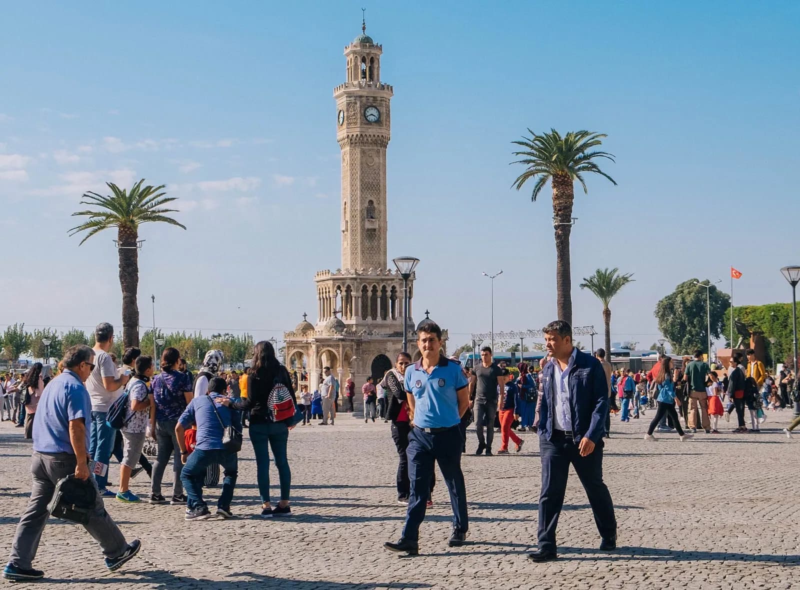 The Clock Tower of İzmir (İzmir Saat Kulesi) rising above Konak Square, surrounded by palm trees and pigeons, with İzmir Bay visible in the background
